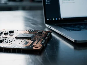 Printplaat met gesoldeerde microchips en kopersporen naast een laptop met code op een stalen werkbank in een engineeringlab.